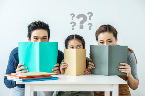 Una giovane madre e un giovane padre asiatici con la loro bambina e un punto interrogativo sopra la testa seduti a una scrivania con un libro in mano e un'espressione divertita sul volto.